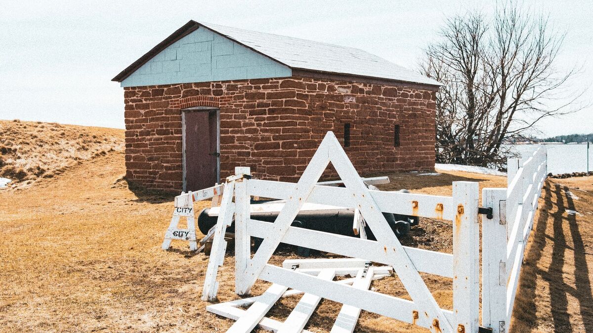 A white board fence with snapped and fallen rails in front of an old stone outbuilding